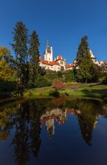 Fototapeta na wymiar Pruhonice, Czech Republic - October 25 2020: Scenic view of romantic castle standing on green hill in a park surrounded with trees. Reflection of the castle in water. sunny autumn day with blue sky.