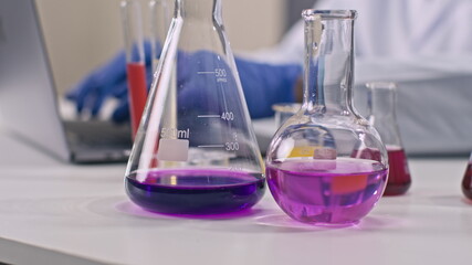Close up view of laboratory glassware with colorful liquids standing on desk in researh medical lab. Scientist works on laptop analysing reaction test tubes, checking chemical formula. Lab equipment