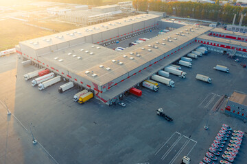Logistics park with warehouse, loading hub and many semi trucks with cargo trailers standing at the ramps for load/unload goods at sunset.  - obrazy, fototapety, plakaty