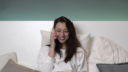 Happy young woman in white pajamas talking on mobile phone and smiling. Attractive sexy brown hair girl chatting in bed before sleep with boyfriend . Social communication concept