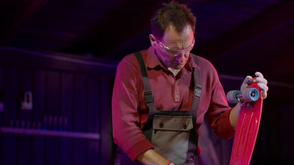 Caucasian artisan in overalls and safety glasses repairs a skateboard or pennyboard in his workshop. Making craft products concept. Men's work. Stylish workshop lighting. Garage for work.