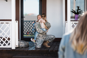 Smiling military service man embracing daughter, while sitting on knee on threshold with blurred...