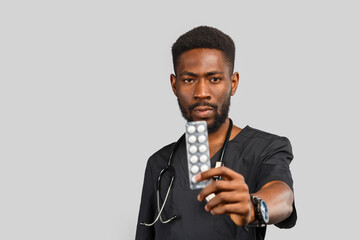handsome african doctor with stethoscope holding pills in hands while standing against gray background