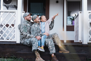Happy military father and mother and daughter pointing with finger, while sitting on house threshold on blurred foreground