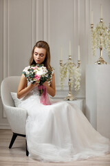 beautiful tender young bride with flower bouquet sitting on armchair in white studio interior