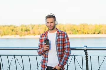 young handsome guy in checkered shirt and modern headphones listening music outdoor while drinking take away coffee, morning