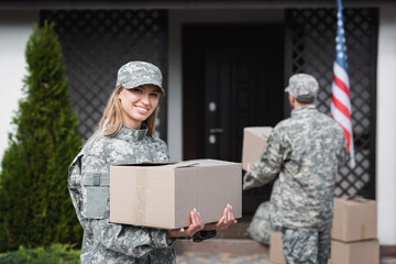 Smiling woman in camouflage holding cardboard box and looking at camera with blurred  military man...
