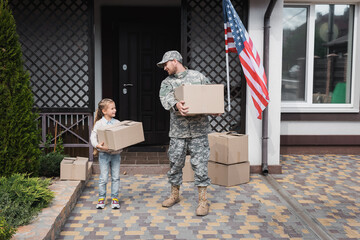 Father in military uniform and daughter holding cardboard boxes near house with American flag