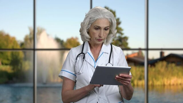 Mature doctor using digital tablet. Female doctor working on tablet pc against windows background with beautiful nature view.