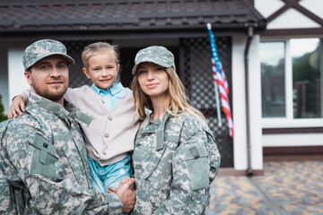 Fototapeta premium Smiling father and mother in military uniforms lifting daughter and looking at camera on blurred background