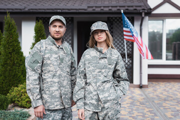 Military couple in uniforms standing together and looking at camera near house