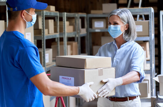 Female manager supervisor wearing face mask preparing fast drop shipping safe delivery giving parcels packages boxes to male courier taking ecommerce orders to deliver standing in warehouse storage.