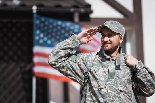 Smiling military man in uniform looking away and saluting with blurred USA flag on background