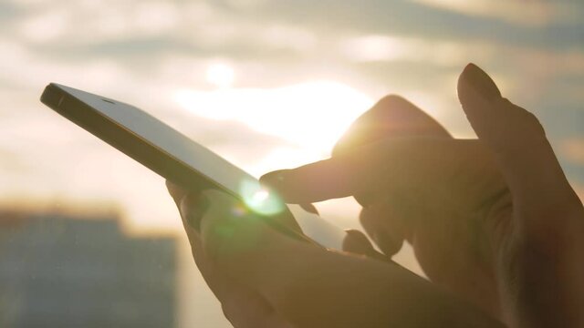 Close up side view: woman hand using smartphone device against warm sunset sky - scrolling and touching. Sun lens flare, golden hour. Relax, entertainment, leisure time and technology concept