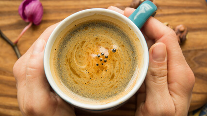 Female hands hold a cup with aromatic coffee. A cup of aromatic coffee stands on a wooden table. Invigorating coffee with delicious milk foam.