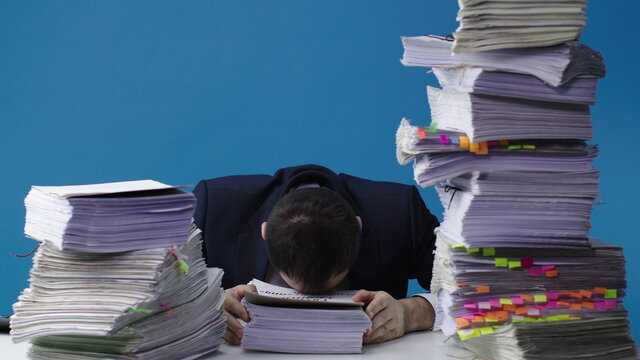 Stressed businessman accountant manager takes stack of unfinished documents from large pile of office papers, in despair lays head on table. Stress at work, overworked, bureaucracy, paperwork concept
