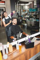 mirror reflection of hairdresser drying hair of client with cosmetic bottles on blurred foreground