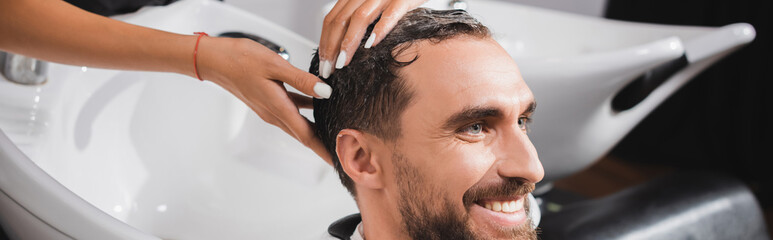 Fototapeta premium partial view of hairstylist washing hair of cheerful bearded man, banner