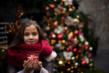 Young pretty pteteen girl  looks at the candle  outdoor at the background decorated christmas tree with lights garland on New Year's Eve holidays. Kid dressed warm jacket, red knitted scarf