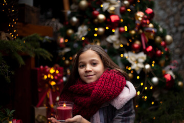 Young pretty pteteen girl  looks at the gift  outdoor at the background decorated christmas tree with lights garland on New Year's Eve holidays. Kid dressed warm jacket, red knitted scarf