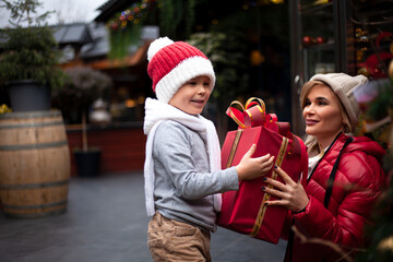 Happy family, young pretty woman with child spend time together outdoor at the background decorated christmas tree with lights garland on New Year's Eve holidays. Kid  holding big gift