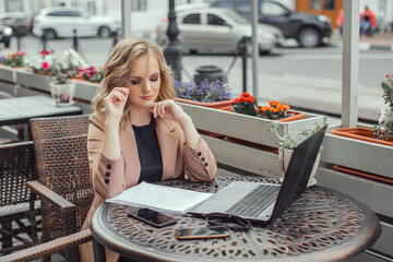 Young business woman works at a computer in a cafe. On the table there is a laptop, phone, notepad. The girl is talking on the phone.