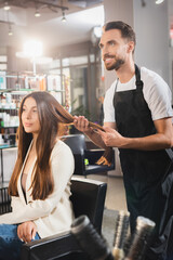 Smiling hairdresser in apron touching hair of young woman, blurred foreground