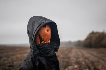 A pumpkin-headed figure Halloween jack-o'-lantern with a scary face in a black hooded robe stands in the  field