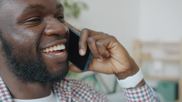 Close-up portrait of cheerful young African American man speaking on mobile phone smiling laughing enjoying communication indoors at home. People and gadgets concept.