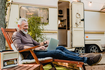 Attractive grey-haired man resting on the wooden deck chair using laptop with caravan van behind - obrazy, fototapety, plakaty
