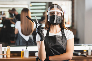 Hairdresser in face shield, latex gloves and apron holding comb and scissors on blurred background