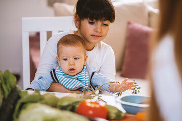Cute girl holding baby boy brother while mother  preparing meal with healthy food 