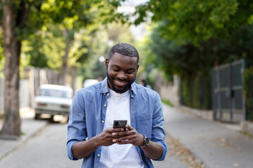Obraz premium Handsome Afro-American man using smartphone and smiling. Happy man using mobile phone apps, texting message, browsing internet, looking at smartphone. Young people working with mobile devices.