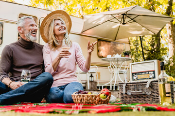 Aged beautiful caucasian couple having picnic and drinking champagne in the yard near the motorhome.Mature blond lady pointing at something