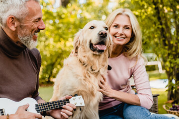 Cheerful mature caucasian couple spending time together with their dog while grey-haired man playing the ukulele in park