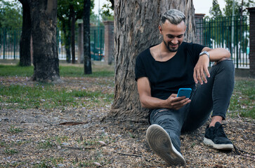 Naklejka na ściany i meble young latin man sitting in the park with a cell phone in his hand smiling 