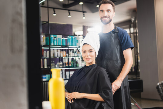 cheerful hairdresser and smiling woman with hair wrapped in towel looking at camera, blurred foreground