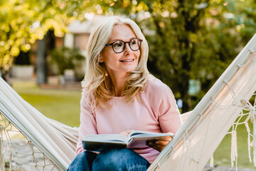 Smiling senior good-looking blond woman wearing glasses while reading in hammock in the summer garden