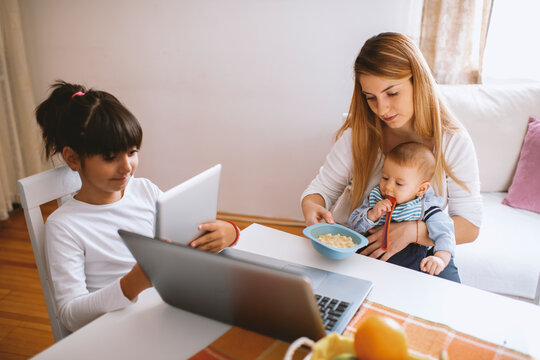 Mother working on laptop, holding baby boy and daughter learning on tablet