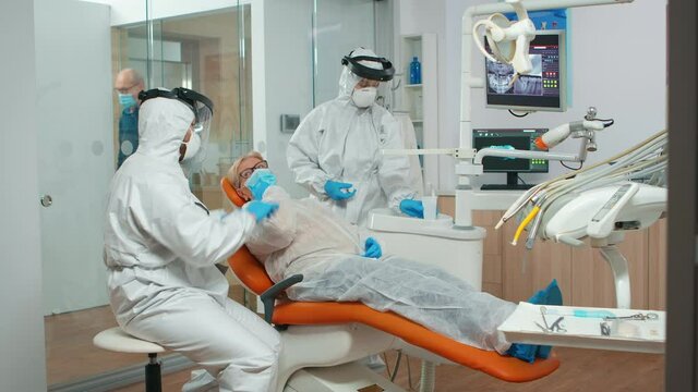 Dental specialist with coverall talking with senior patient before examination during pandemic covid-19. Concept of new normal dentist visit in coronavirus outbreak wearing protective suit and face
