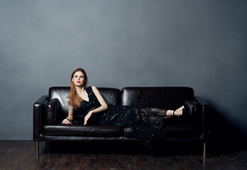 An elegant lady in a black dress lies on a leather sofa indoors in full growth