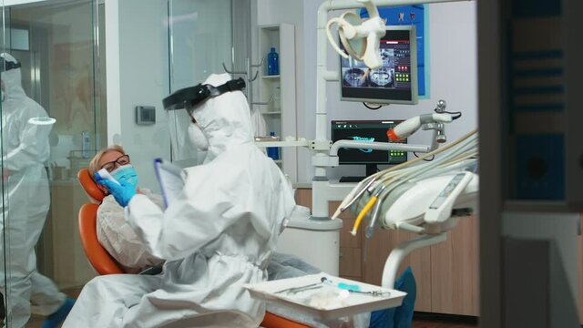 Nusre with face shield measuring old woman temperature before dental examination during global pandemic. Concept of new normal dentist visit in coronavirus outbreak wearing protective suit and face