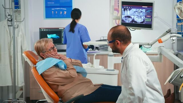 Retired woman having symptoms of gums pain holding hand on cheek while talking with doctor. Elderly patient explaining dental problem to doctor indicating mouth while nurse preparing sterile tools.