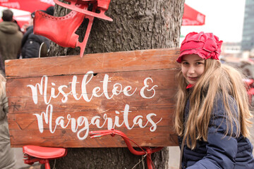 Cute young girl with red hat stand beside christmas wooden sign mistletoe and red bike. Tree with sign and skates. Christmas time 