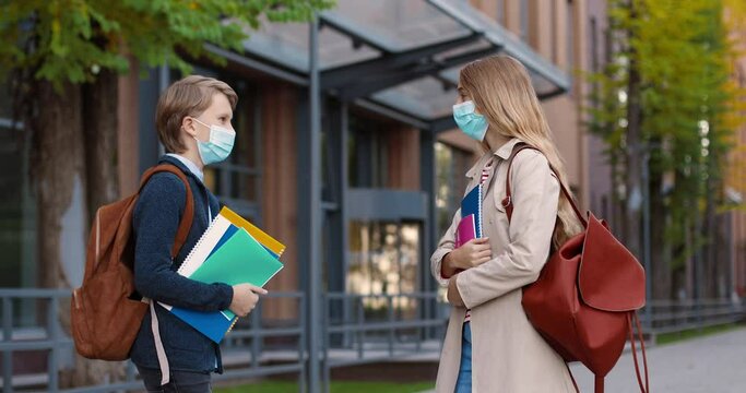 Portrait of teen boy in mask with backpack meeting near school with girl classmate and talking. Caucasian schoolgirl greeting with elbow with friend with copybooks and notebooks outdoors Study concept