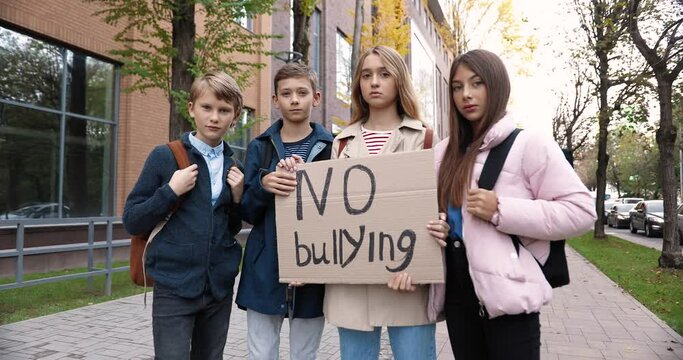 Portrait of cute serious teen schoolkids standing outdoors near school with "No bullying" sign. Boys and girls college students holding poster against mocking and intimidation. Children concept