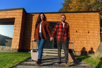 Handsome bearded man and pretty stylish woman go out from house with suitcases and going to trip.