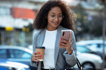 Pretty young business woman using her smart phone while drinking coffee walking down the street.
