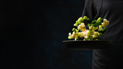 Professional chef in black uniform throws up frying mix of cabbage and broccoli above the pan on dark blue background. Backstage of cooking meal. Frozen motion. Food banner concept.