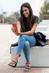 Pretty young woman making video call with smartphone sitting on a bench in the street.